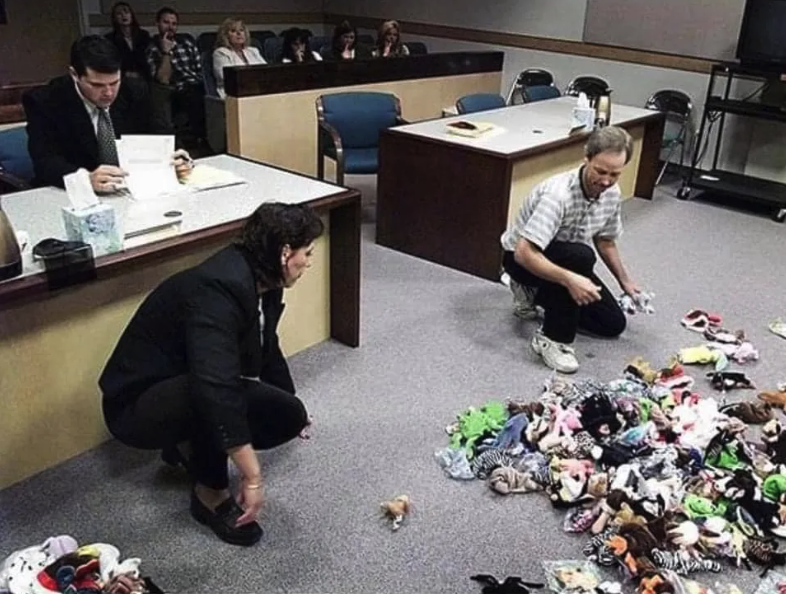 A photograph of two people crouched down on the floor in a courtroom, sorting through an enormous pile of tiny stuffed animals that surely will be worth a fortune in 26 years.